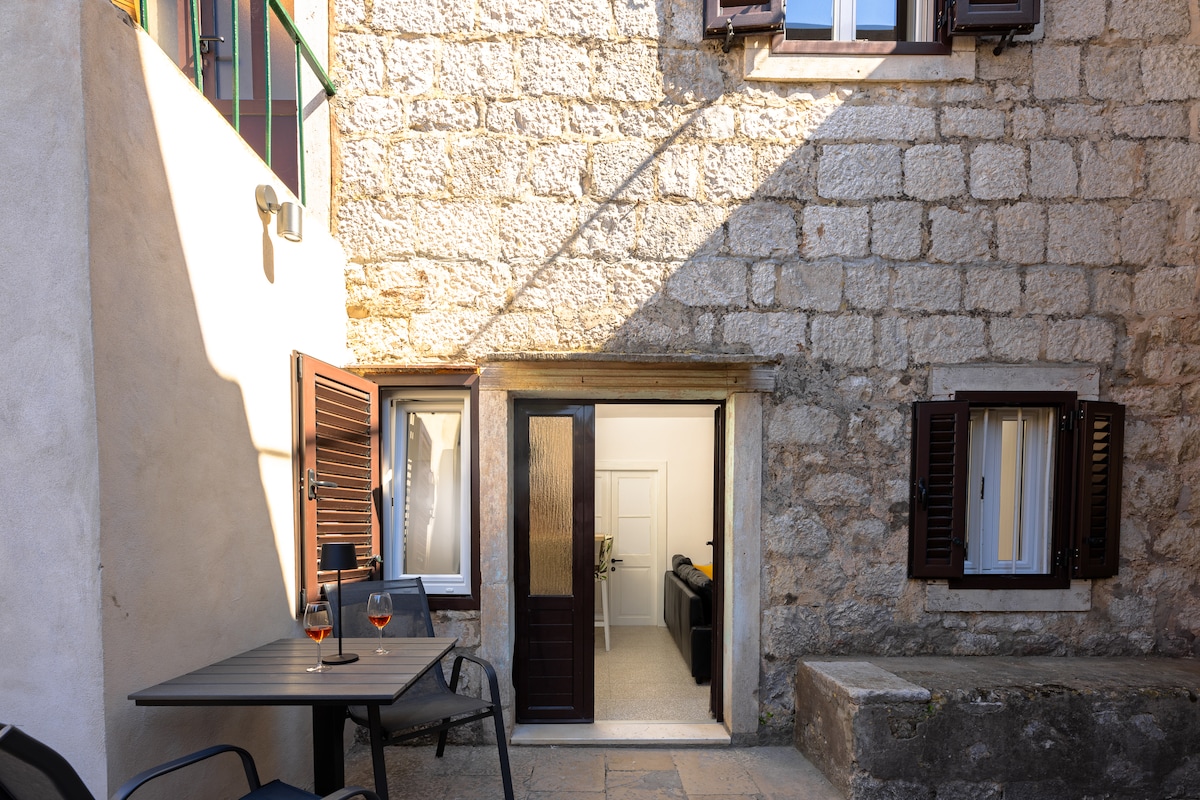 An outdoor seating area is situated near the entrance, featuring a small table and two black chairs. The stone walls are illuminated by sunlight, highlighting the open doors leading into the interior space. Window shutters are partially open, allowing natural light to flow inside.