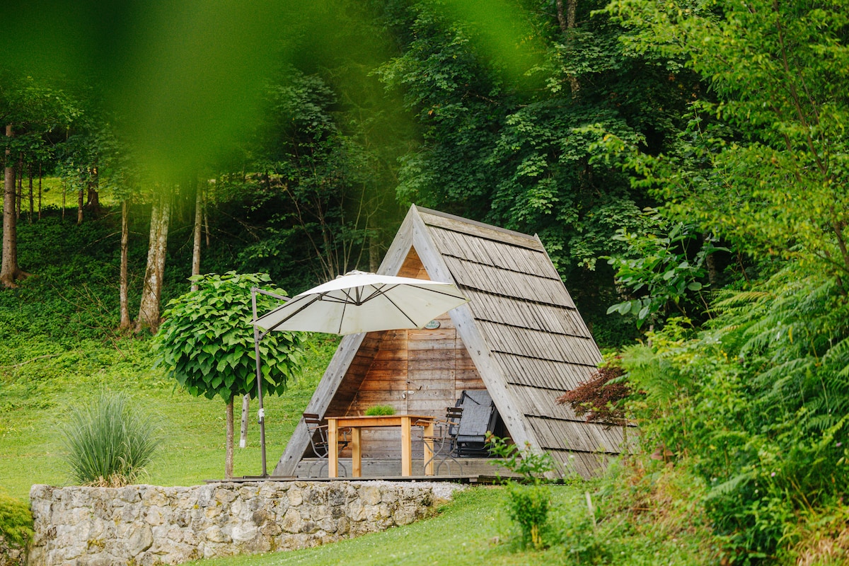An A-frame cottage is nestled in a lush green landscape, featuring a wooden exterior and a sloped roof. A shaded outdoor area is equipped with a table and chairs. Surrounding foliage creates a serene setting, enhancing the connection to nature.