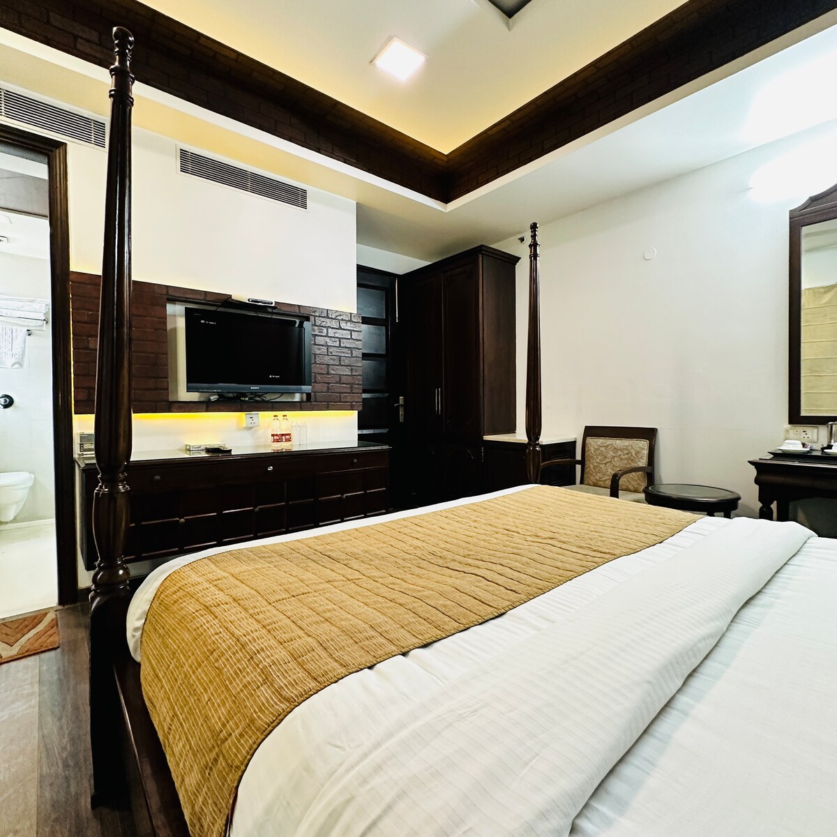 The sleeping area features a spacious bed draped in a light blanket, with dark wood accents throughout the room. A wooden dresser holds a television, and a full-length mirror is positioned beside a chair. Soft lighting creates a cozy ambiance.