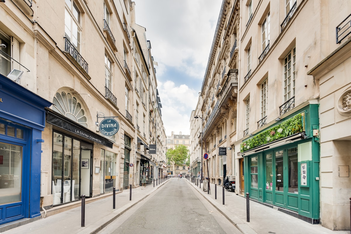 A charming street in a historic Parisian neighborhood is lined with elegant buildings featuring balconies and decorative facades. Shops with inviting storefronts can be seen on both sides, while a bright green tree adds a touch of nature to the scene. A clear sky above enhances the overall ambiance.