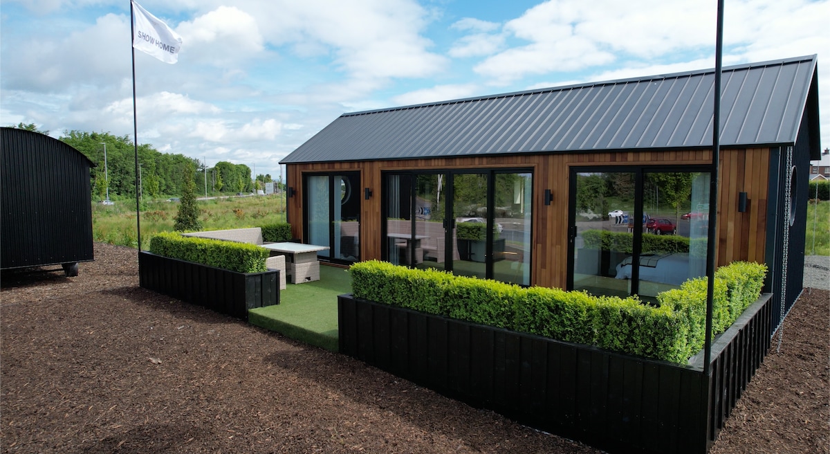 A modern wooden cabin is depicted, featuring large glass windows and a sleek metal roof. Surrounding green hedges are neatly trimmed, while a paved area with outdoor furniture is visible in front of the cabin. The landscape includes clear skies and distant trees.