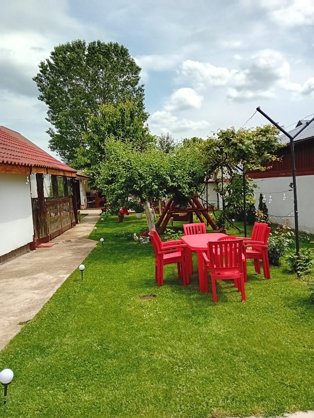 A grassy outdoor area features a red table surrounded by matching chairs, providing a place for relaxation. Nearby, a swing set is visible under a lush green tree, enhancing the natural setting. Soft white lights illuminate the path leading toward the main building.