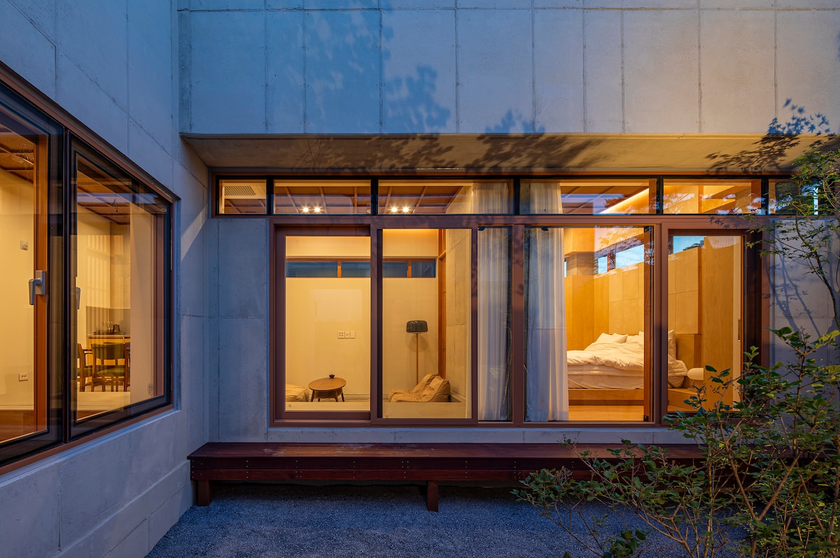 A serene exterior view of a modern accommodation is presented, featuring large windows that allow a glimpse into the interior. The warm glow of light from inside highlights a spacious bed and minimalistic furnishings. A small outdoor area is accessible, framed by muted landscaping.