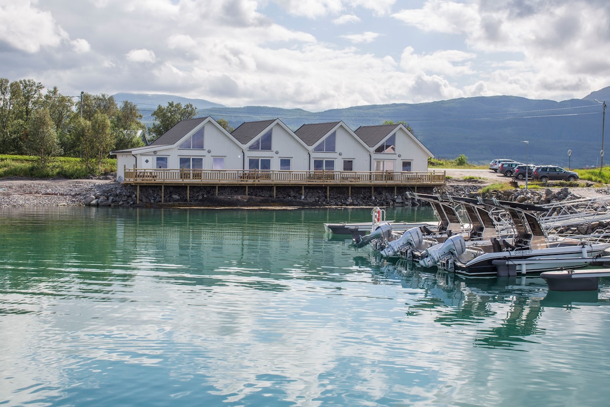 The image displays a row of modern sea lodges situated along the water's edge, featuring a wooden deck. A marina with several boats is visible in the foreground, and lush greenery and distant mountains frame the backdrop under a partly cloudy sky.