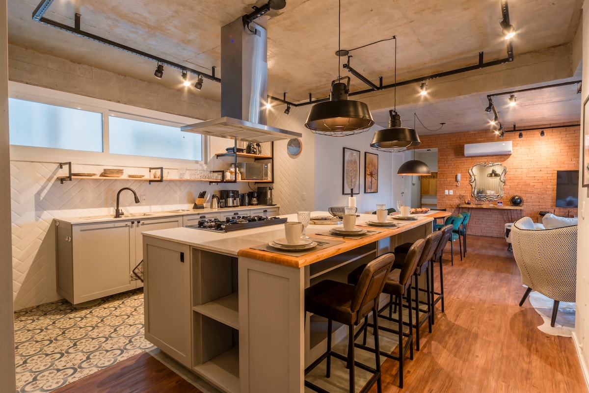 A gourmet kitchen is highlighted by a spacious island with seating for six, complemented by modern pendant lighting. The kitchen features sleek cabinetry, a gas stovetop, and stylish backsplash tiles, while the adjacent area showcases an inviting rustic brick wall.