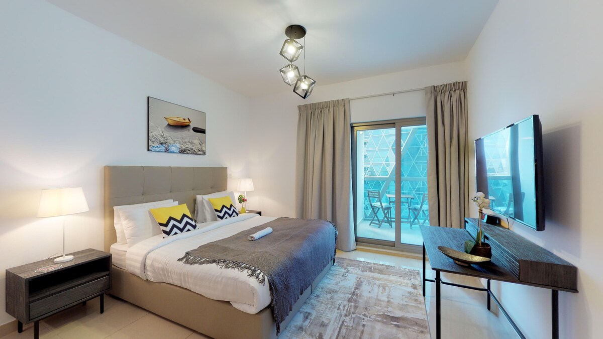 A spacious bedroom features a comfortable bed adorned with decorative pillows. Soft lighting is provided by stylish lamps on either side. A television is mounted on the wall opposite the bed. Large glass doors lead to a private balcony with outdoor seating.