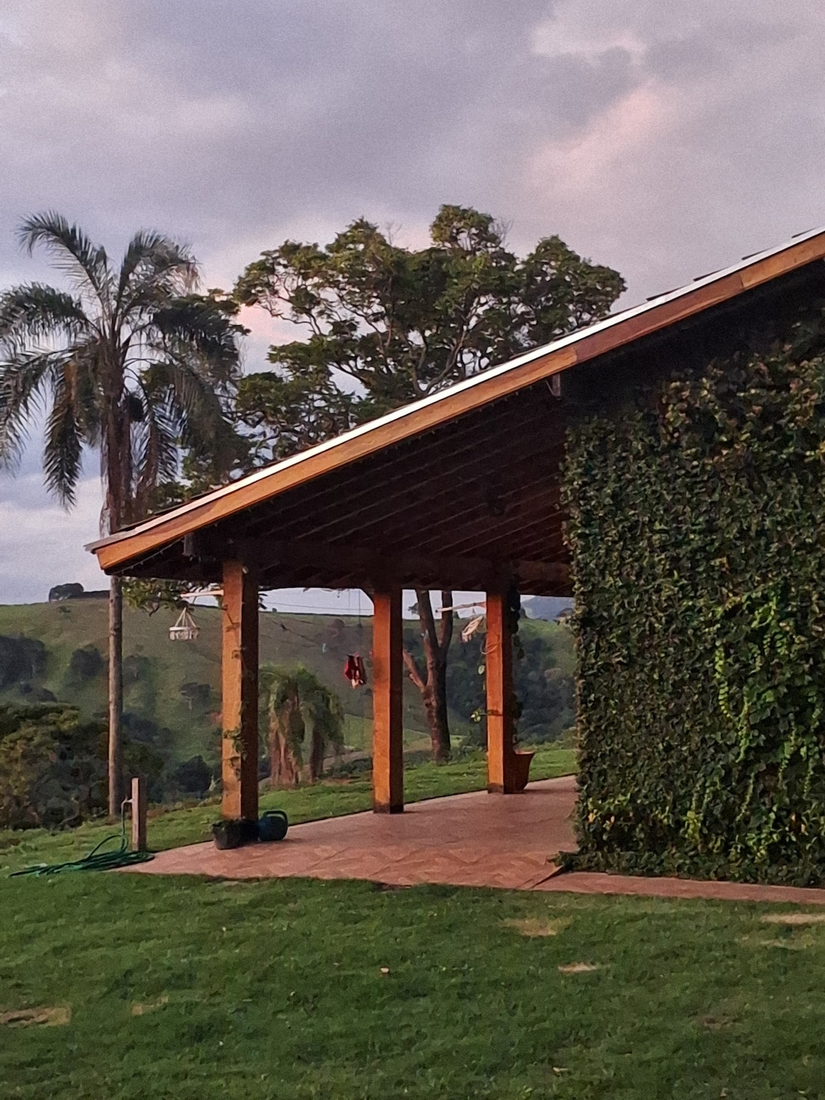 A wooden patio with a sloped roof is framed by lush greenery and tall trees. The structure showcases wooden pillars and a rustic facade, blending with the surrounding natural landscape. Soft evening light enhances the tranquil atmosphere, inviting connection with the outdoors.
