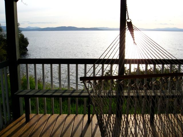 A hammock is suspended on a deck overlooking the expansive waters of Lake Champlain. Soft light reflects off the water's surface, while distant hills create a serene backdrop against the evening sky.