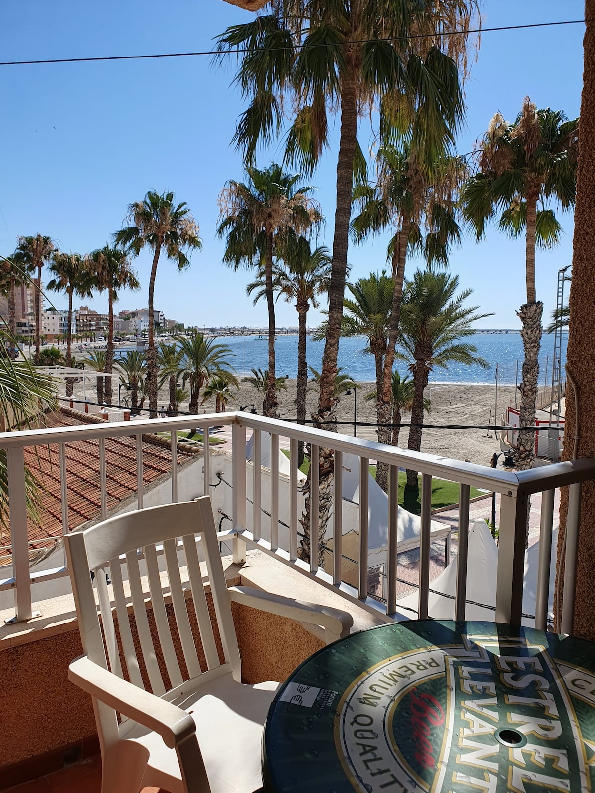 A balcony view features palm trees swaying in the breeze, overlooking a sandy beach and the sea. A white chair and a round table are positioned against the railing, providing a relaxing spot to enjoy the scenery.
