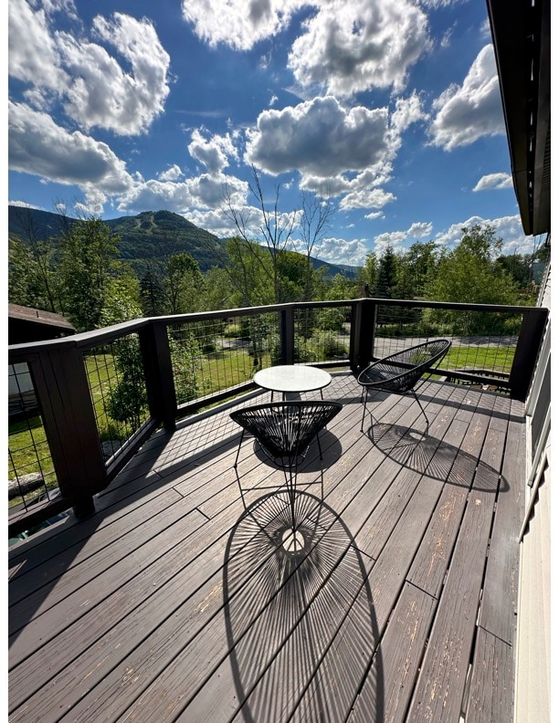 An outdoor deck area features a round table and two modern chairs, providing a place to relax and enjoy the surrounding mountain views. The wood flooring adds a natural element, with shadows created by the furniture contrasting against the bright sky filled with fluffy clouds.