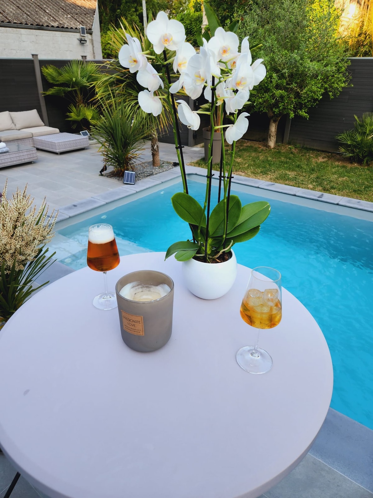 A round table is set by a clear swimming pool, featuring two glasses of beverages and a decorative candle. A white orchid in a pot adds a touch of greenery. Lush plants surround the area, creating a serene outdoor space.