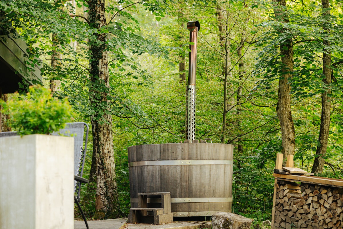 A wooden hot tub is surrounded by lush greenery, with a stone surface leading to it. A small step platform provides access, and a stack of firewood sits nearby, enhancing the natural setting. The scene conveys a sense of tranquility amidst the forest.