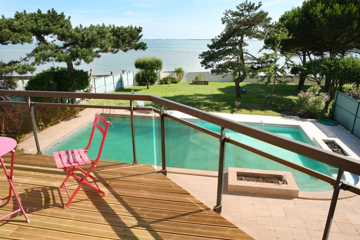 A private swimming pool is framed by a wooden deck, with a pink chair inviting relaxation. Lush greenery surrounds the area, leading to a picturesque view of the bay. The serene landscape is complemented by clear blue skies and calm water.