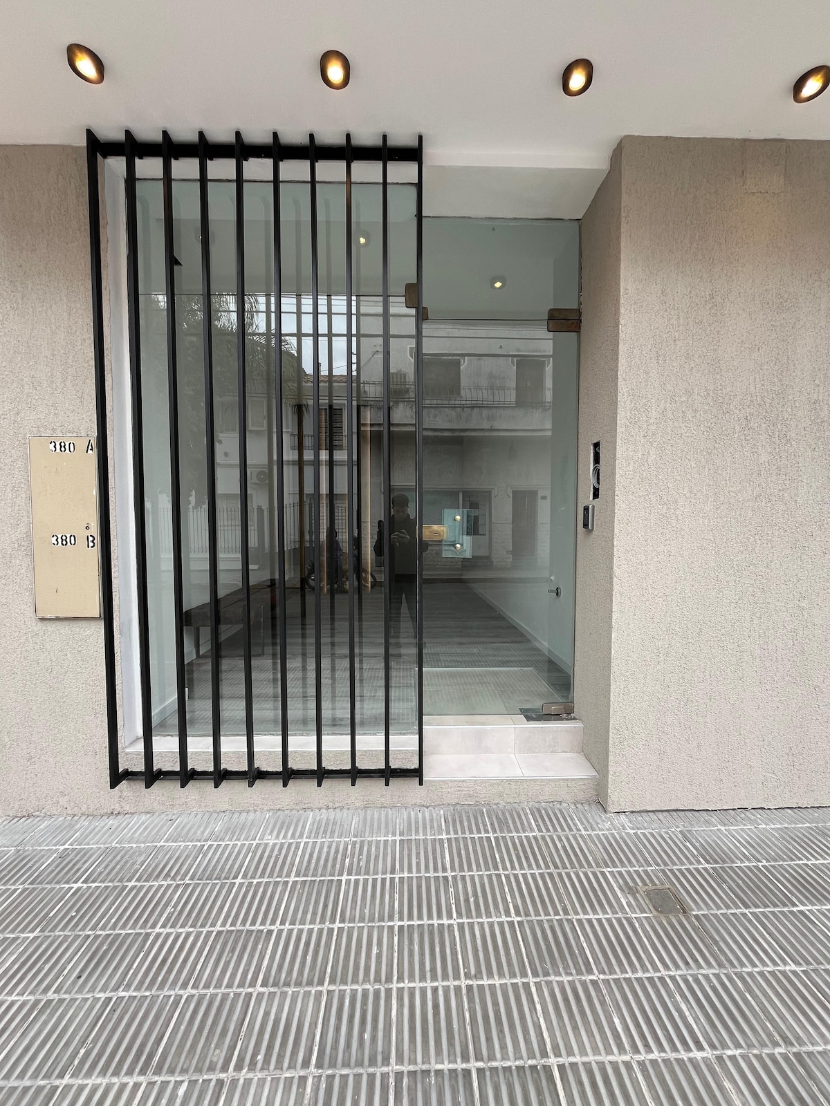 The entrance features a clear glass door framed by sleek black metal bars. The floor is lined with modern tiled patterns, leading into a well-lit lobby area. Neutral-toned walls provide a contemporary feel, complemented by discreet overhead lighting.