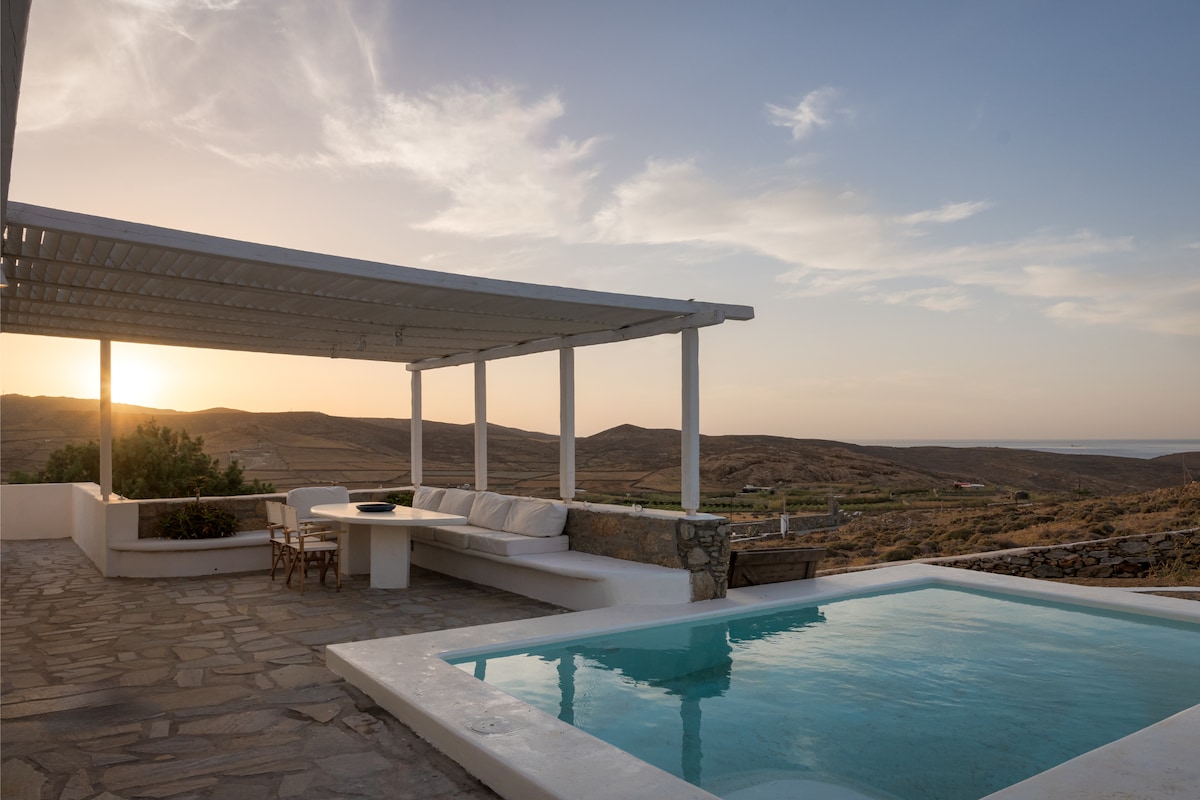 An outdoor terrace is showcased with a spacious seating area and a dining table under a white pergola. A private pool reflects the soft evening light, complemented by expansive views of the surrounding mountains and a gentle sunset.