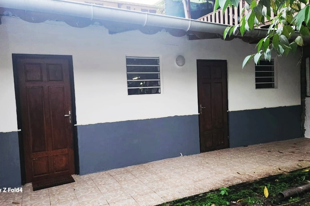 Two entrance doors are positioned side by side, each featuring a simple wooden design. Light-colored walls contrast with the dark lower section, while small windows allow natural light to filter in. A tiled pathway leads to the doors, framed by greenery.