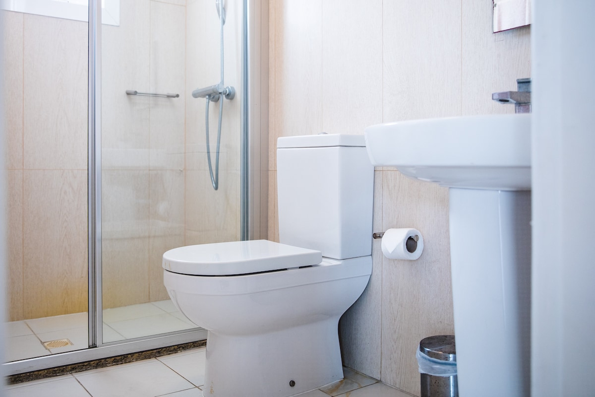 A clean bathroom is displayed, featuring a modern toilet and a shower area with transparent glass doors. Light tiles complement the neutral wall tones, while a small waste bin is positioned near the toilet. Basic amenities such as toilet paper are readily available.