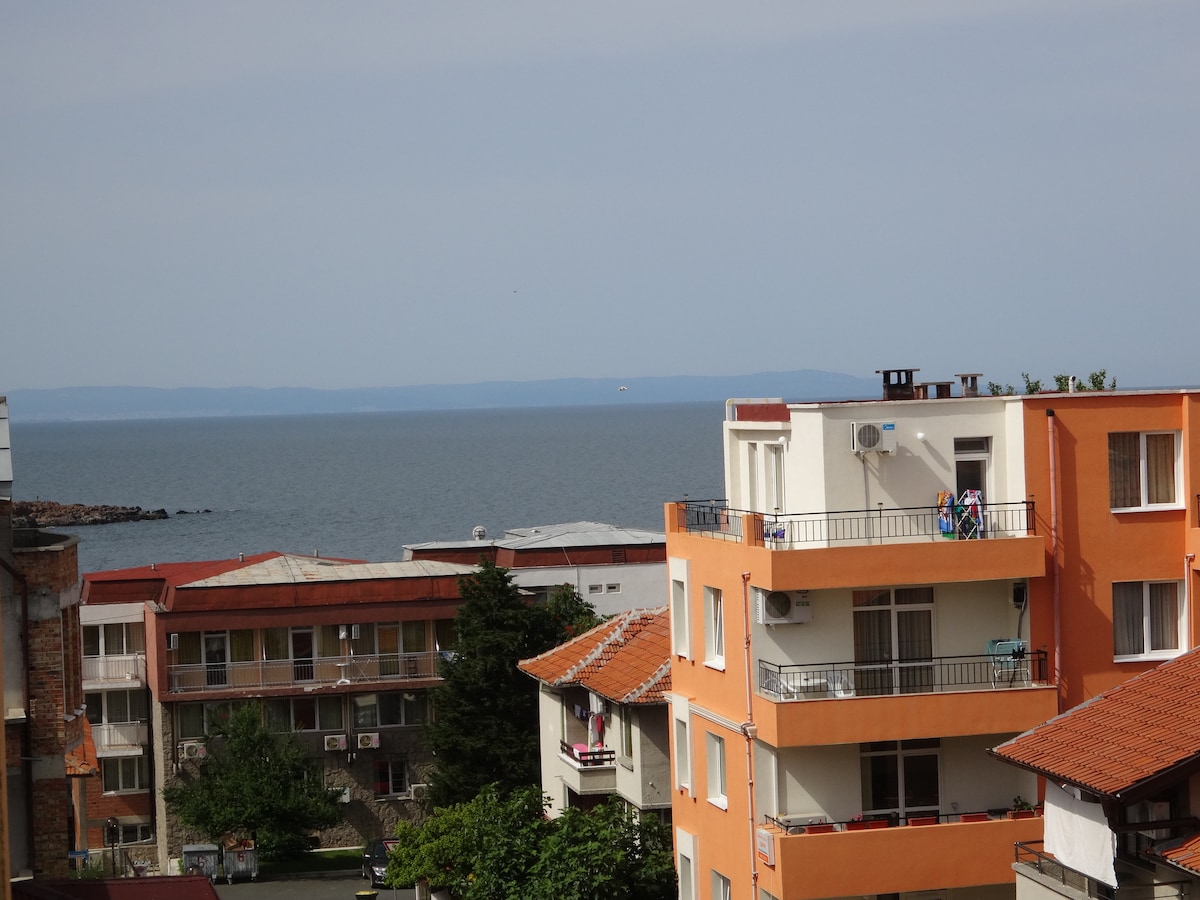 The image captures a view of the sea from a distance, with buildings in the foreground. The calm water extends to the horizon, where it meets a partly cloudy sky. Various residential structures with balconies and terracotta roofs are visible, offering a glimpse of local architecture.