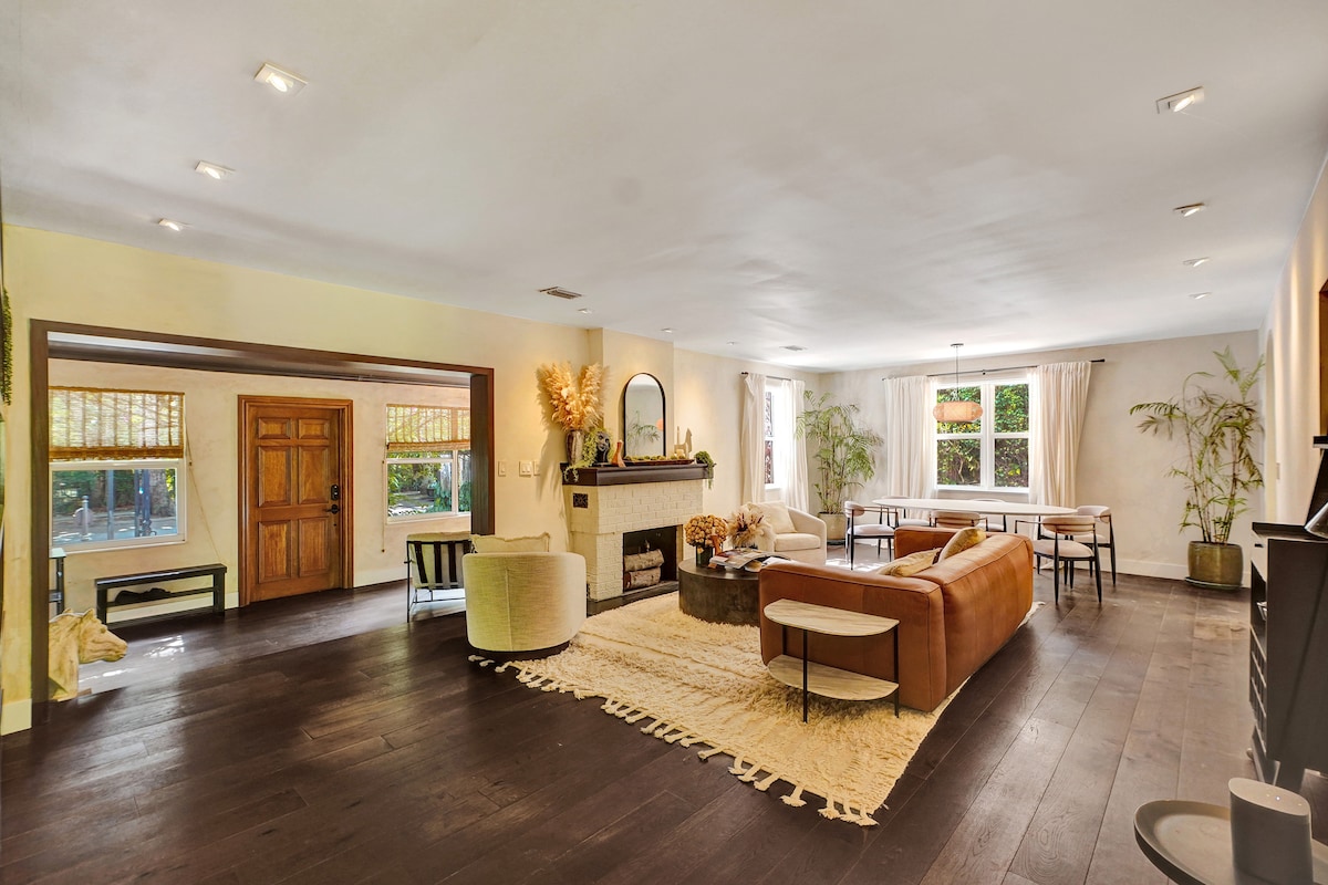 A spacious living area features large windows allowing natural light to fill the room. A textured area rug lies on hardwood floors, complemented by a modern beige sofa and a leather couch. A decorative fireplace is centered along one wall, surrounded by plants and stylish furnishings.