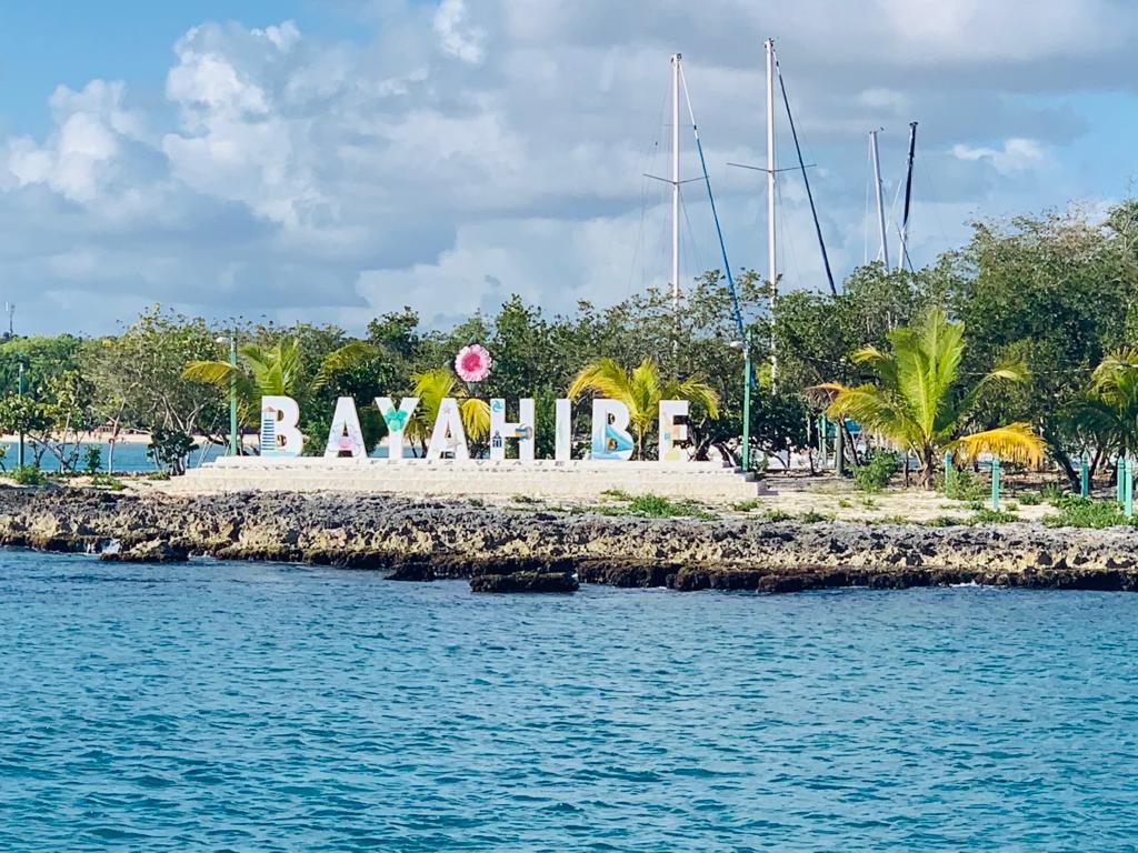 The image showcases the Bayahibe sign prominently positioned along the shore, surrounded by greenery and palm trees. The water reflects the vibrant colors of the sign under a partly cloudy sky, while sailboats are visible in the background, emphasizing the coastal setting.