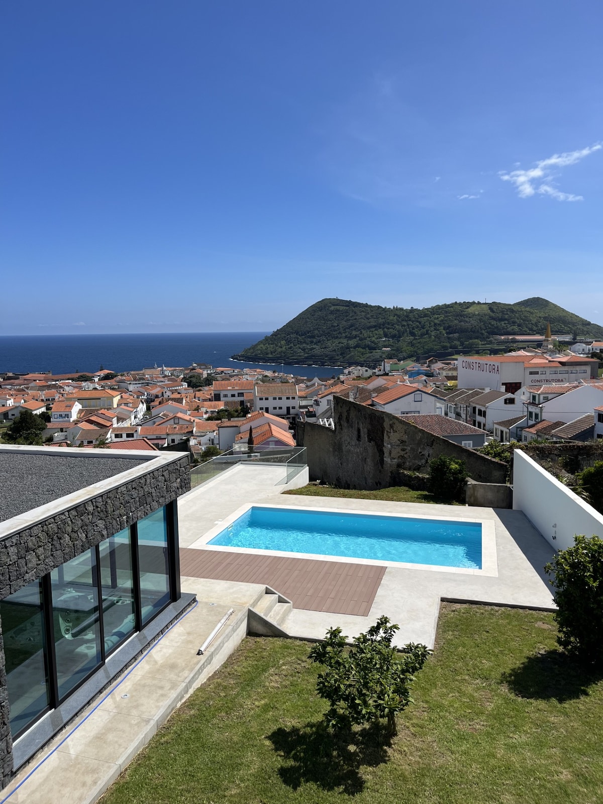 A panoramic view showcases a contemporary villa with a swimming pool nestled within a landscaped area. The ocean sparkles in the distance, framed by the scenic hills and rooftops of Angra do Heroísmo, offering a serene backdrop to the outdoor space.