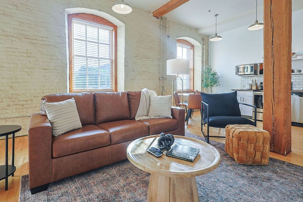 A cozy living area features a brown leather sofa and a comfortable black armchair, complemented by a round wooden coffee table. Large windows allow natural light to fill the space, highlighting the exposed brick walls and wood beams that add character to the room.