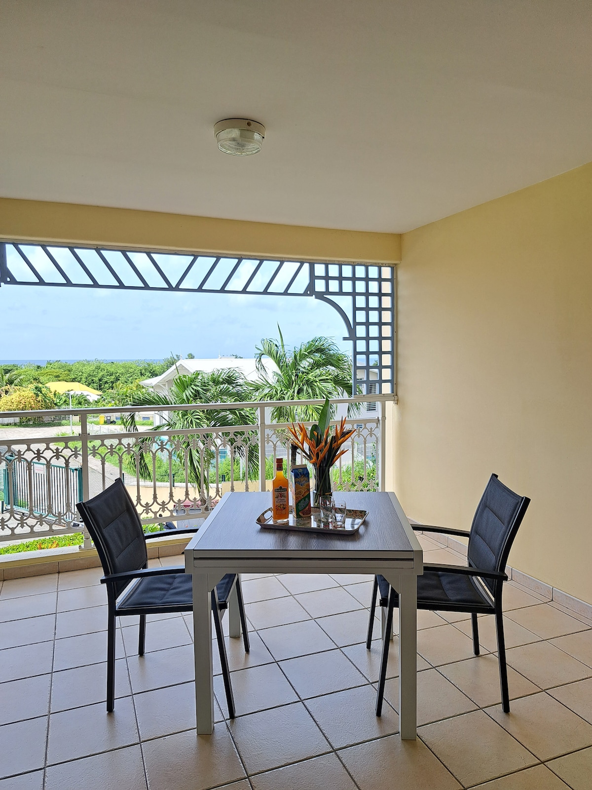 A dining table is positioned on a terrace, accompanied by two black chairs. A tray with drinks and fresh fruit adorns the table. Beyond the railing, a scenic view of greenery and the coast is visible, with bright skies above.