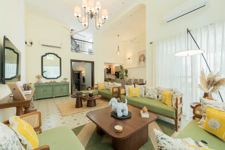 A spacious and stylish living area with high ceilings, curated art, and warm natural light streaming through the windows. The chic chandelier, handcrafted furniture, and plush seating make it an inviting space for lounging and gatherings.