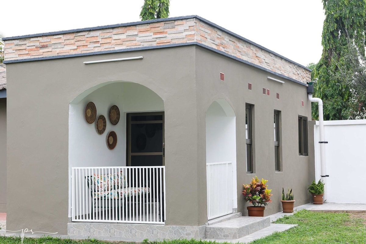 A modern studio is presented with a smooth grey exterior and a decorative stone accent along the top. A small covered patio features comfortable seating, while potted plants add natural beauty. Large windows offer ample light, enhancing the inviting character of the space.