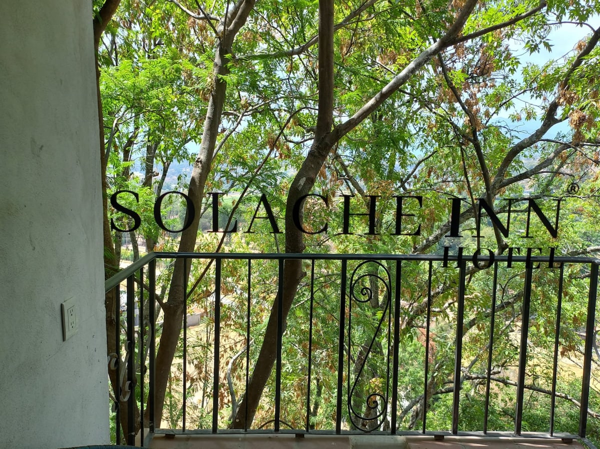A balcony offers a view overlooking lush trees, creating a serene atmosphere. The railing is designed with intricate wrought iron details, complementing the natural surroundings, while soft light filters through the foliage.