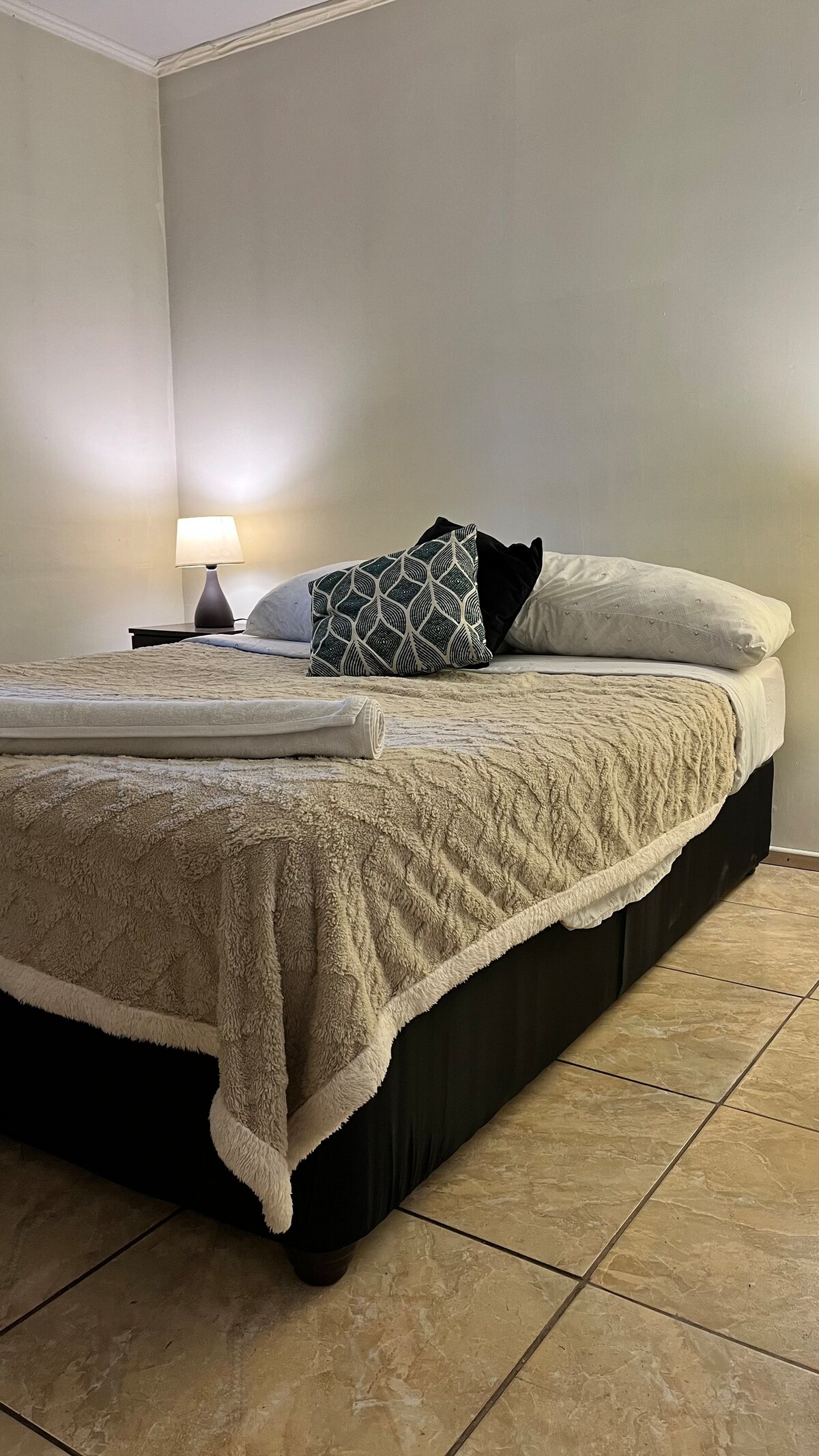 A comfortable sleeping area features a spacious bed dressed with a beige quilt and soft pillows. A bedside table holds a lamp providing soft illumination, and the warm tones of the walls enhance the tranquil atmosphere. Light-colored tiles add to the room's calming effect.
