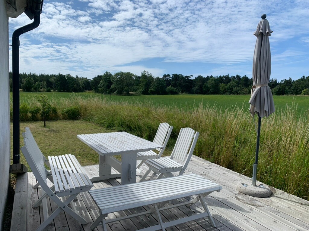 An outdoor deck features a white table surrounded by five matching chairs, complemented by a sun umbrella. The setting offers views of expansive green fields and is framed by tall grass under a blue sky with scattered clouds.