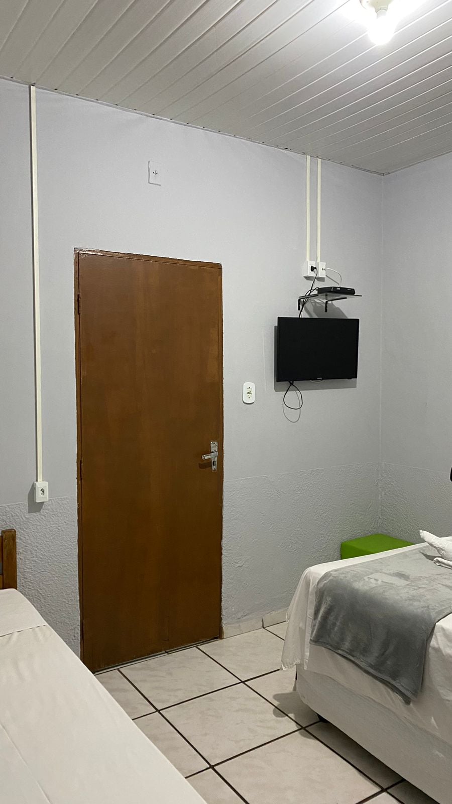 A simple guest room is presented with two beds positioned against the walls. A door leads to the hallway, while a television is mounted on the wall opposite the beds. Light-colored walls and tiled flooring contribute to a clean and functional appearance.