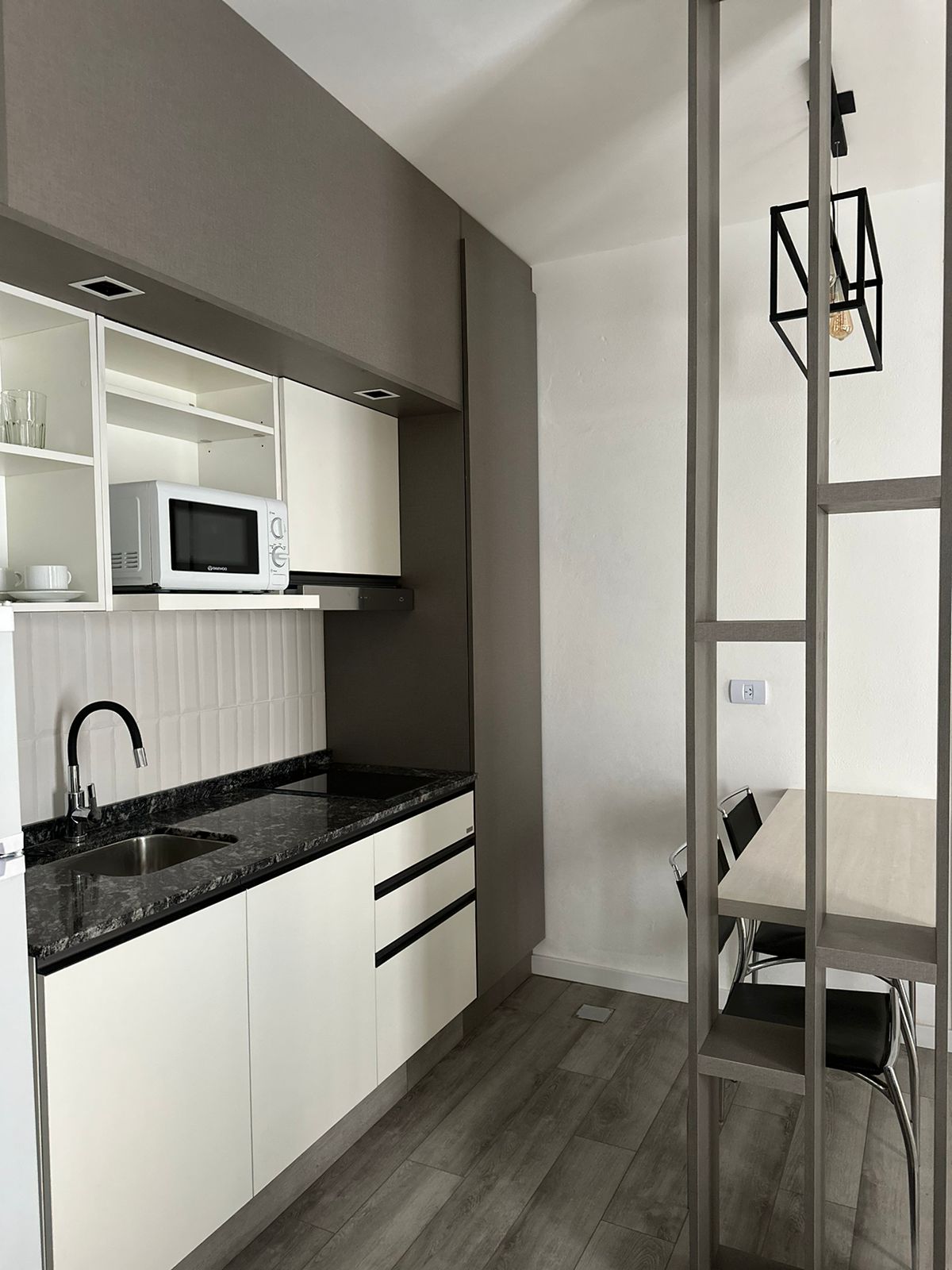 A modern kitchen area features light-colored cabinetry with a dark countertop. A microwave sits above a sink, and open shelving displays glassware. A dining space includes a light wood table paired with black chairs, with a geometric light fixture hanging above.