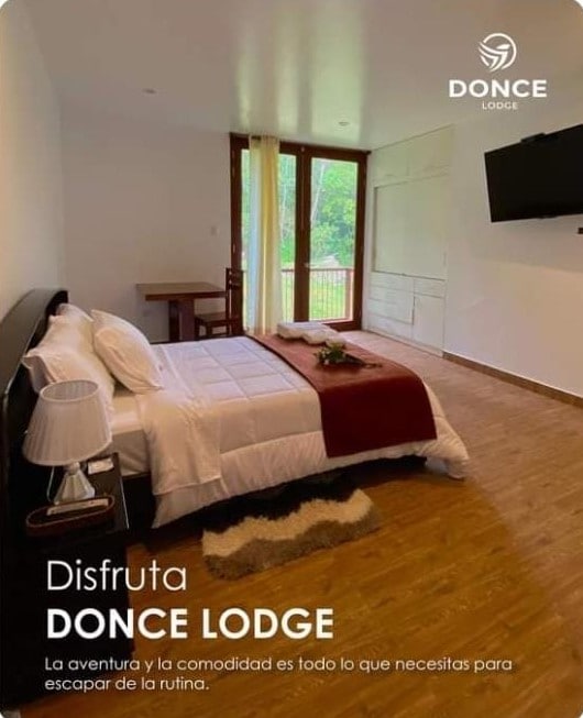 A comfortable bedroom features a spacious bed dressed in white linens, accompanied by a dark wooden headboard. Natural light fills the room through large windows facing lush greenery. A flat-screen television is mounted on the wall, providing modern convenience.