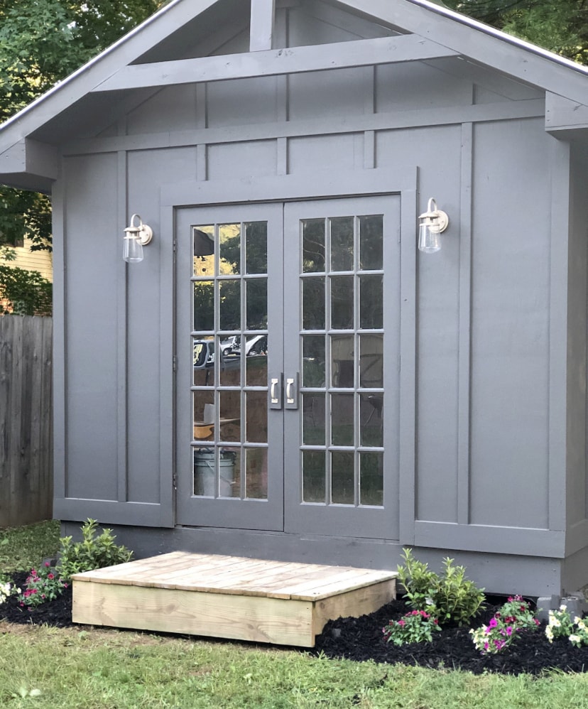 A gray wooden structure features large glass double doors, providing a view of the interior. Flanking the entrance, small green plants add color to the outdoor space, complemented by a wooden step leading up to the door. A privacy fence forms a backdrop.