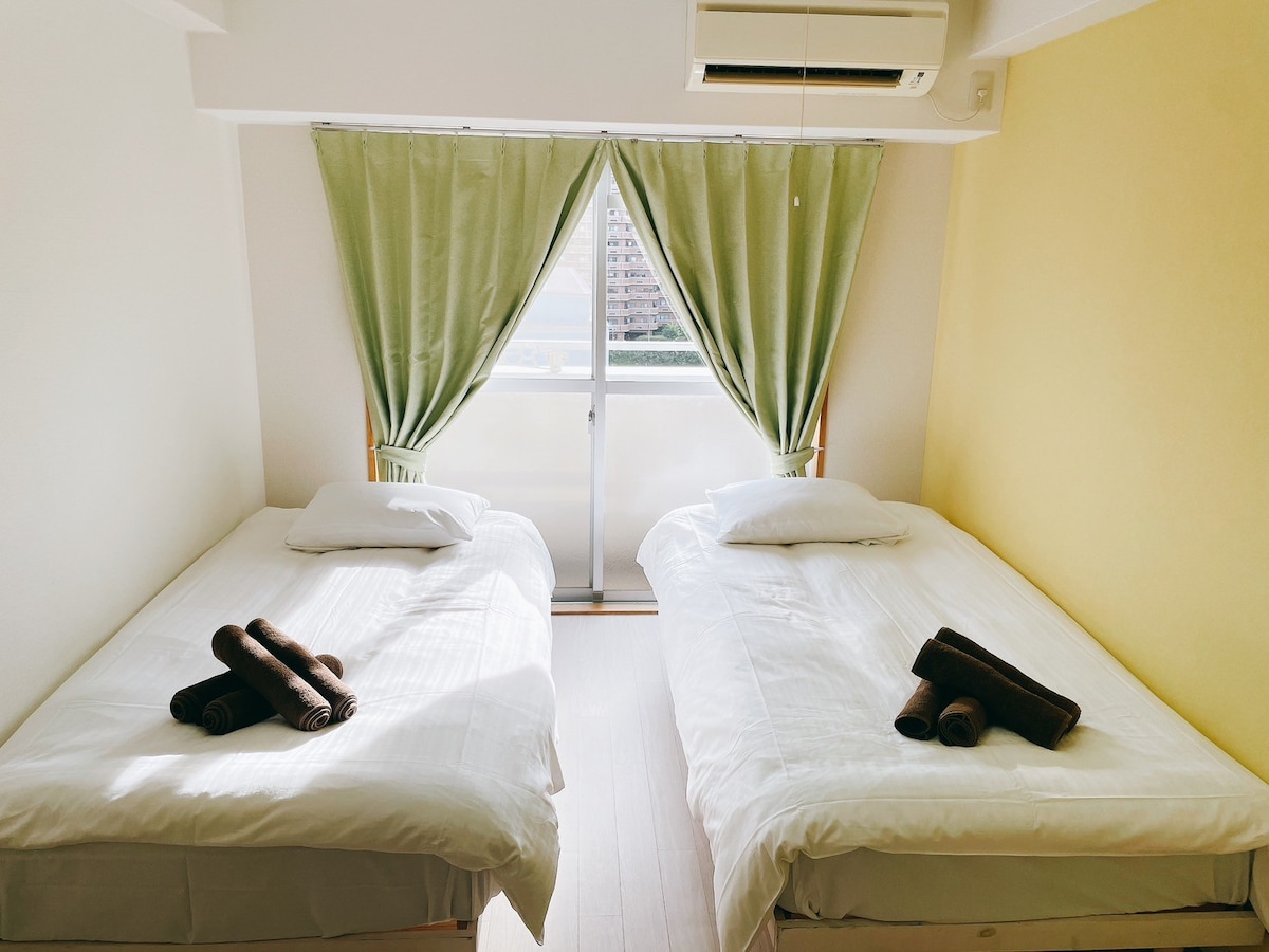 The bedroom features two single beds with crisp white bedding, positioned beside a large window adorned with light green curtains. Towels are neatly rolled and placed on each bed, complementing the calming yellow wall opposite.