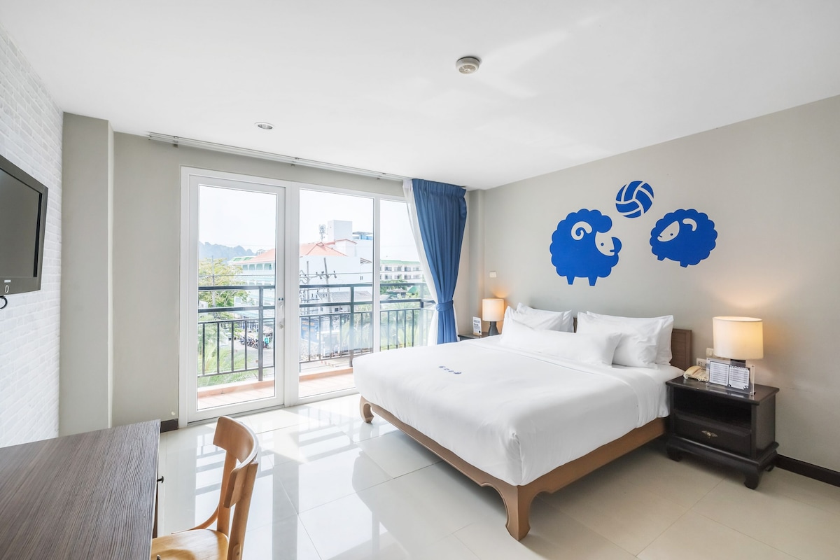 A well-lit guest room features a spacious bed facing large sliding glass doors that lead to a balcony. A bright blue sheep mural adds a playful touch to the decor. A desk and chair are positioned near the window, complemented by bedside lamps and a flat-screen TV.