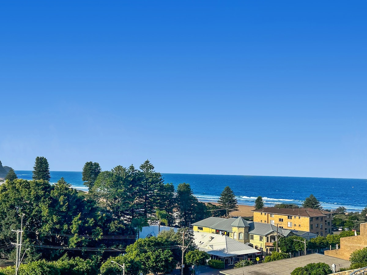 A clear coastal view showcases a serene beach, framed by lush greenery and several houses in the foreground. The ocean's gentle waves are visible under a bright blue sky, creating a tranquil seaside atmosphere.