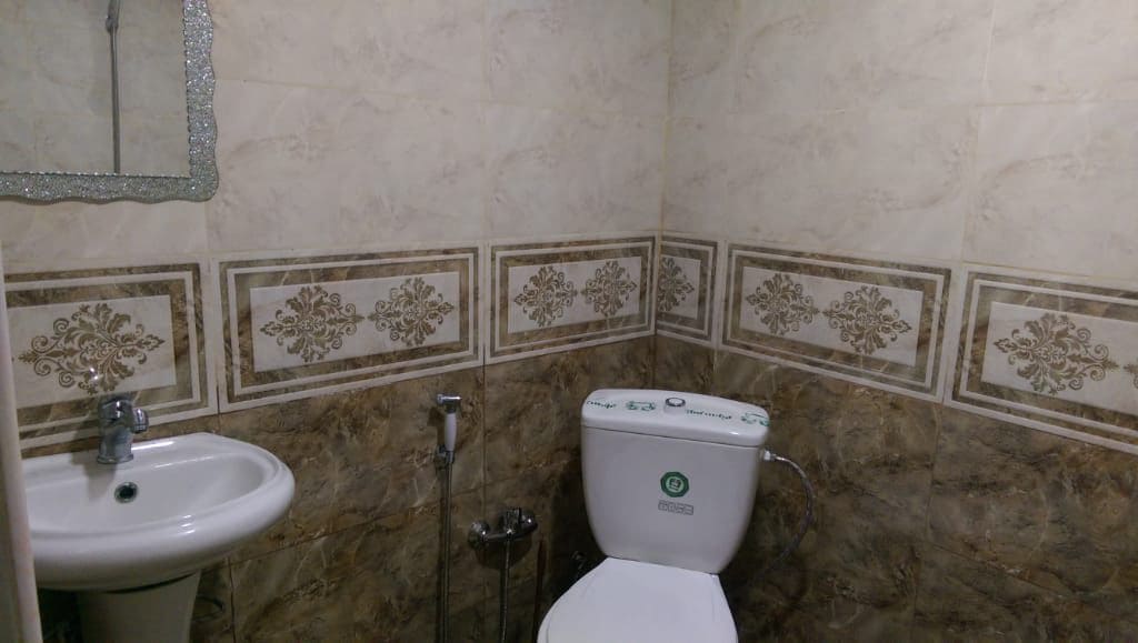 A compact bathroom features beige and gold patterned tiles covering the walls. A white toilet is positioned beside a small chrome bidet. A pedestal sink is mounted beneath a decorative mirror, providing a functional and clean space.