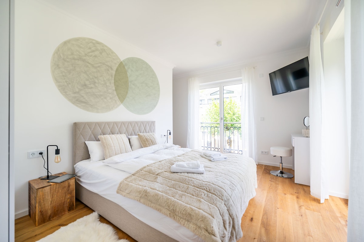A modern bedroom features a spacious bed with neutral linens and a textured throw. An accent wall with circular designs complements the decor. Natural light fills the room through balcony doors leading to a view of greenery, while a wall-mounted television adds convenience.