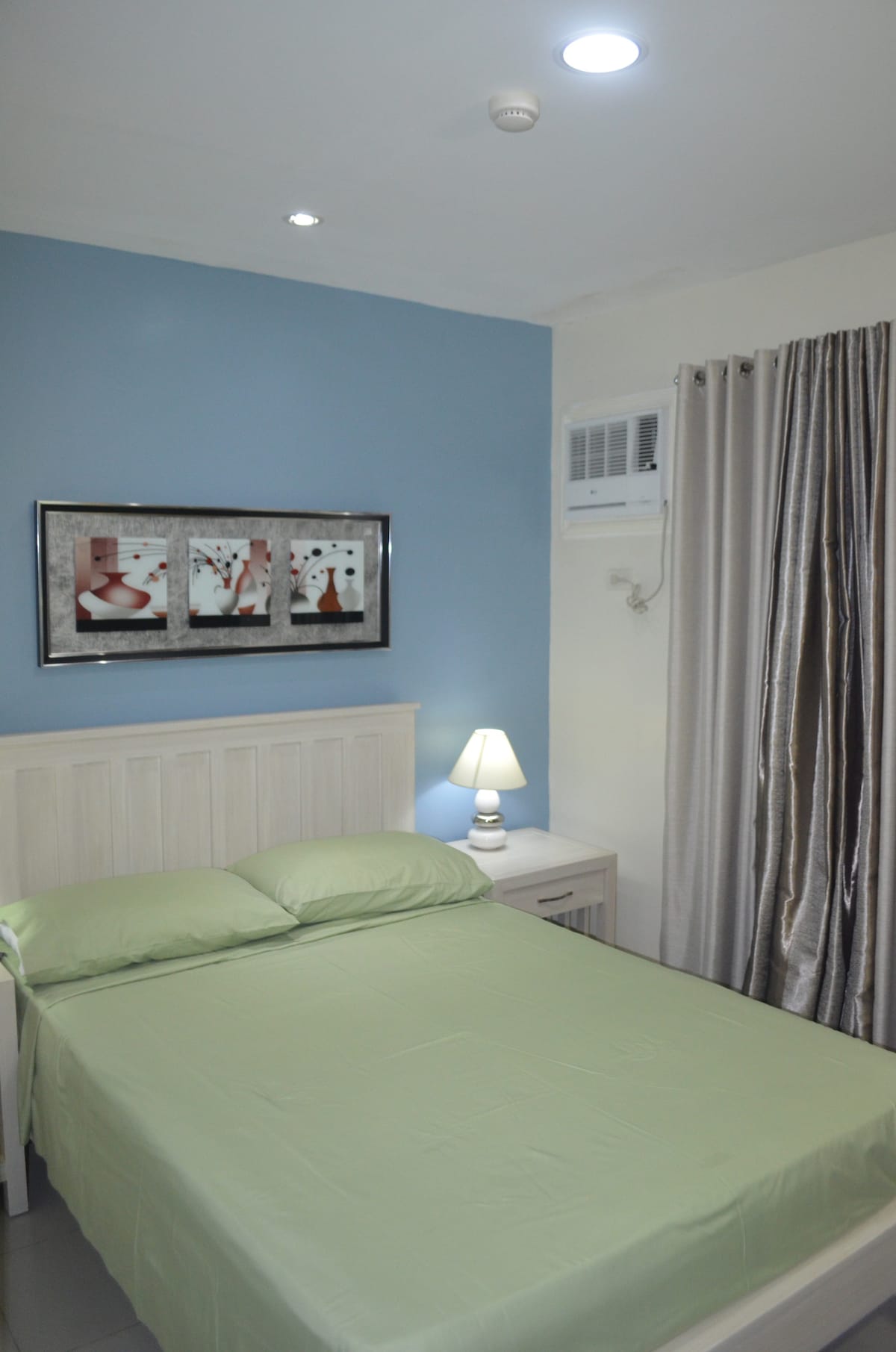A serene bedroom features a soft green bedspread on a spacious bed, accompanied by a neutral-colored headboard. A bedside lamp emits soft light, and a framed art piece adorns the wall. Light drapes complement the room's palette, and a window air conditioning unit is visible.