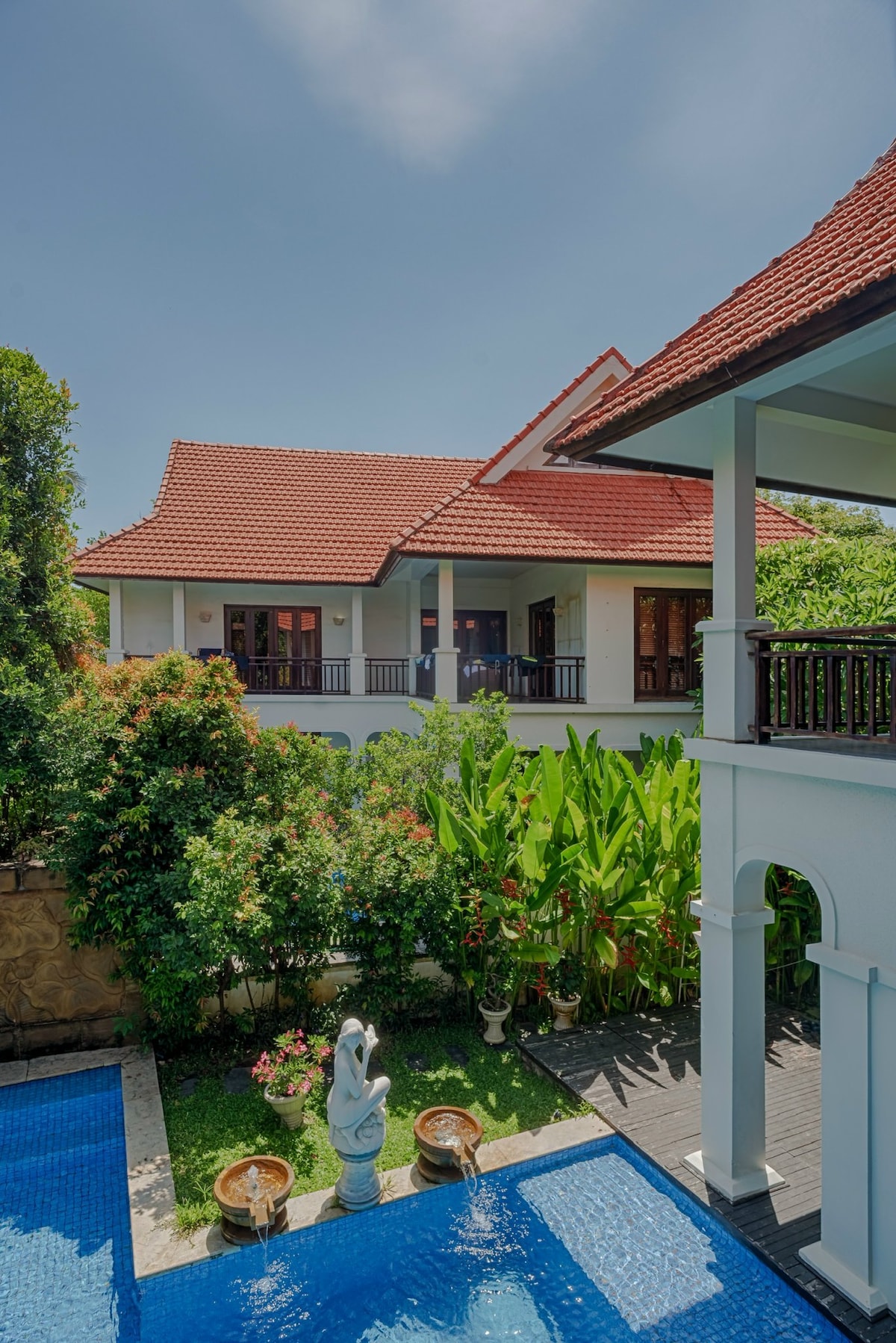 A tranquil outdoor space features a clear blue pool surrounded by lush greenery. The two-story house with a red-tiled roof is visible in the background, along with a stone statue and flower arrangements enhancing the garden area.