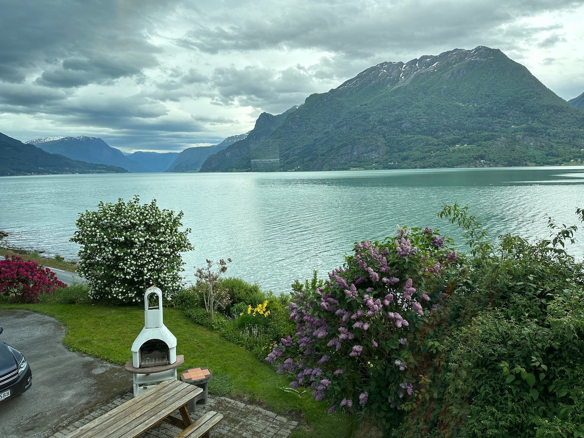 A serene view of the fjord is framed by lush greenery and colorful flowering shrubs. A stone outdoor fireplace is positioned near a wooden picnic table, inviting relaxation. The distant mountains reflect the clouds above, creating a calm and tranquil atmosphere.