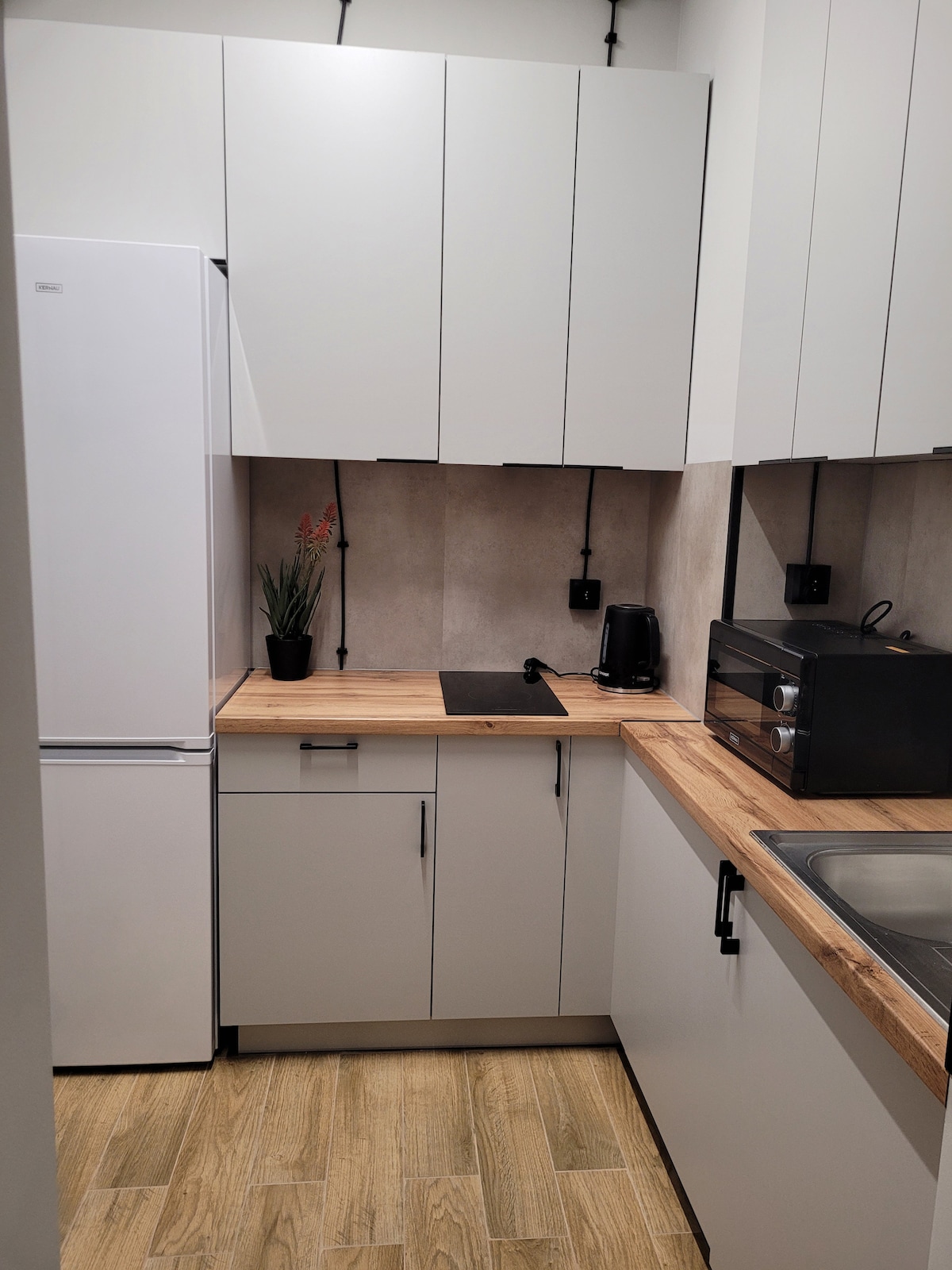 A compact kitchen area features minimalist cabinetry in white with wooden countertops. Essential appliances include a refrigerator, a coffee maker, and a toaster, all positioned for convenience. A small potted plant adds a touch of greenery to the space, enhancing the clean and functional design.