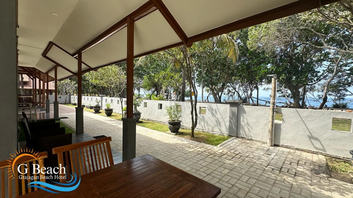 A pathway lined with greenery is visible, leading to the beach area. Outdoor seating is provided near wooden structures, with potted plants positioned along the walkway. The ocean is seen in the distance, framed by trees and natural light filtering through the leaves.