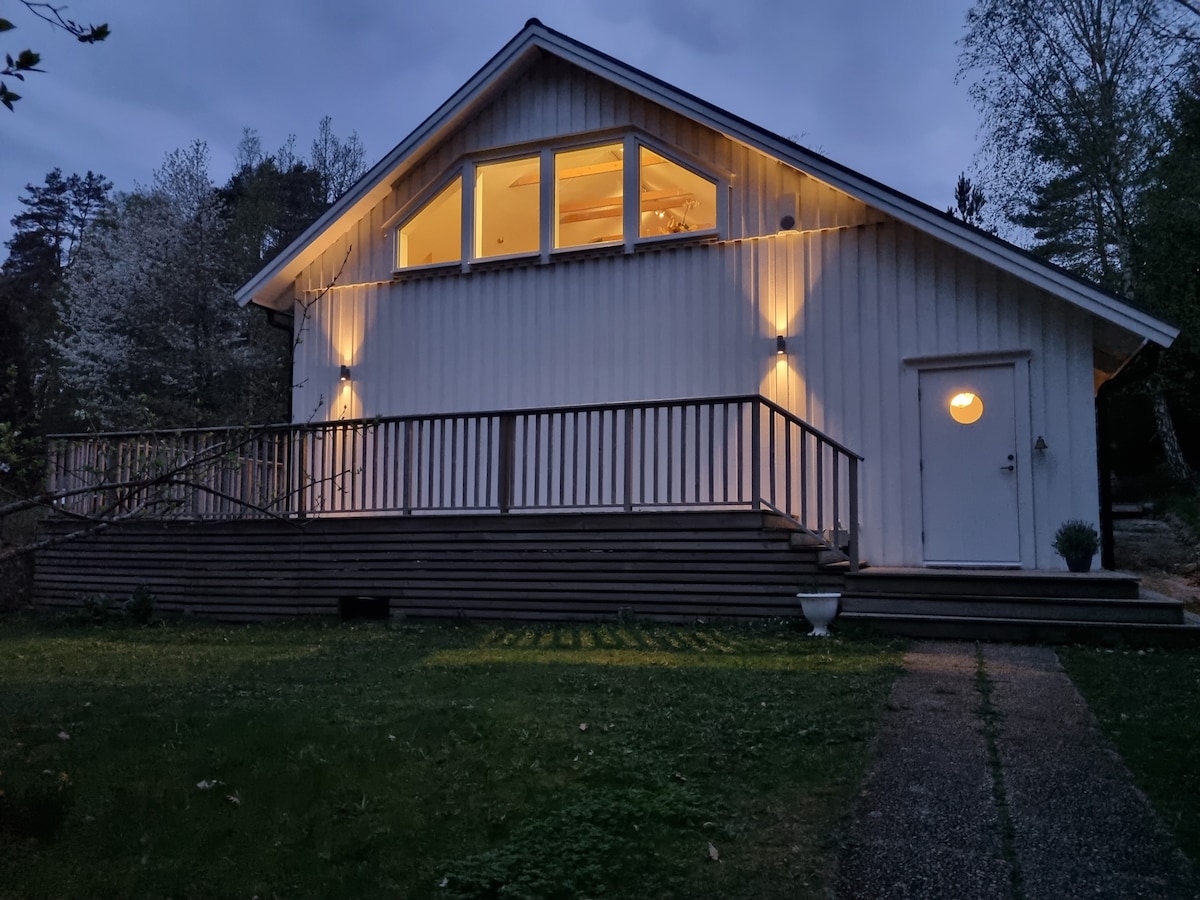 A modern house with a sloped roof is illuminated by warm lighting in the evening. Large windows are located on the upper level, reflecting the soft glow. A wooden deck extends along the front, leading to a path bordered by grass and trees.