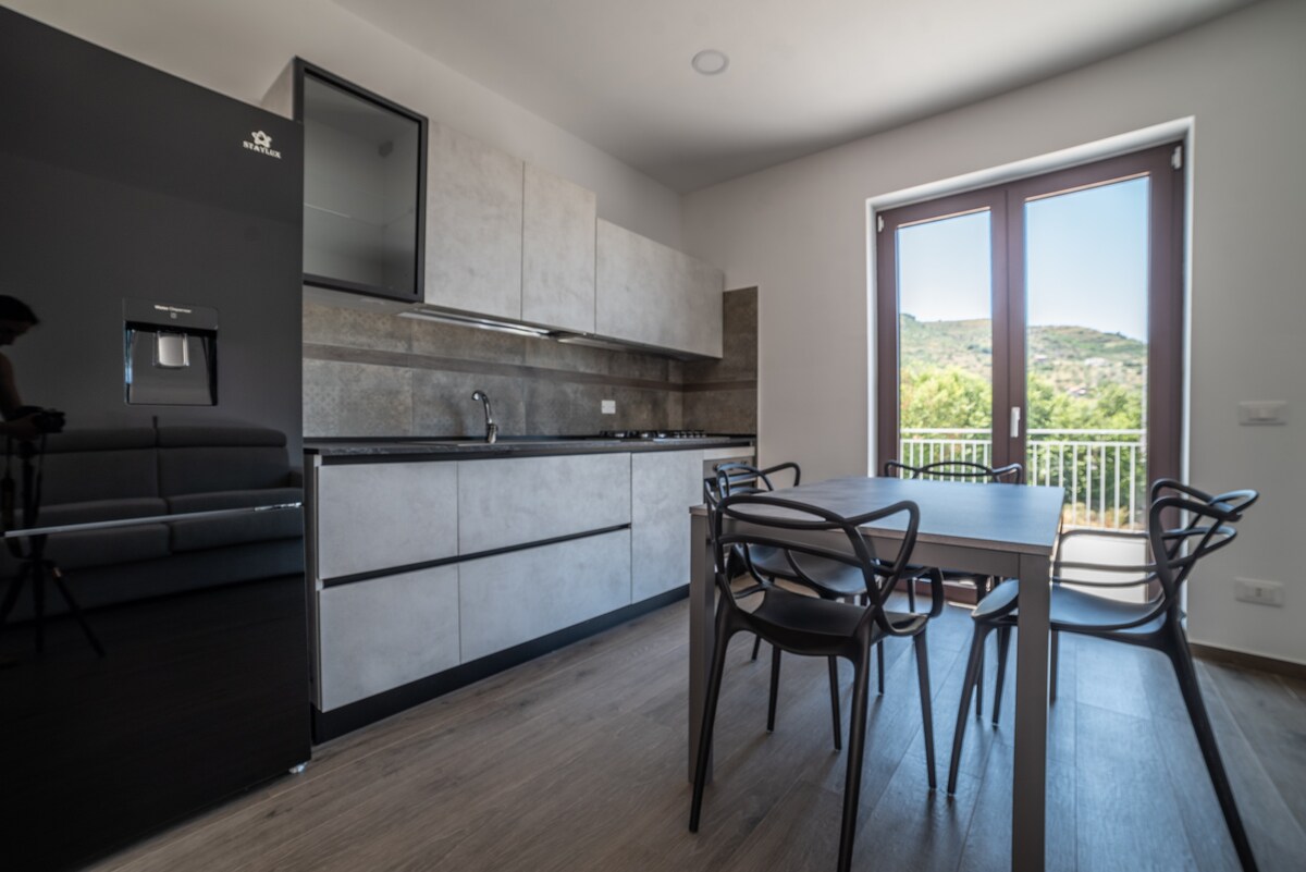 A modern kitchenette is shown with a sleek design featuring light cabinetry and dark countertops. A dining table with four black chairs is positioned near the glass doors that open to a scenic view of the countryside, allowing natural light to fill the space.