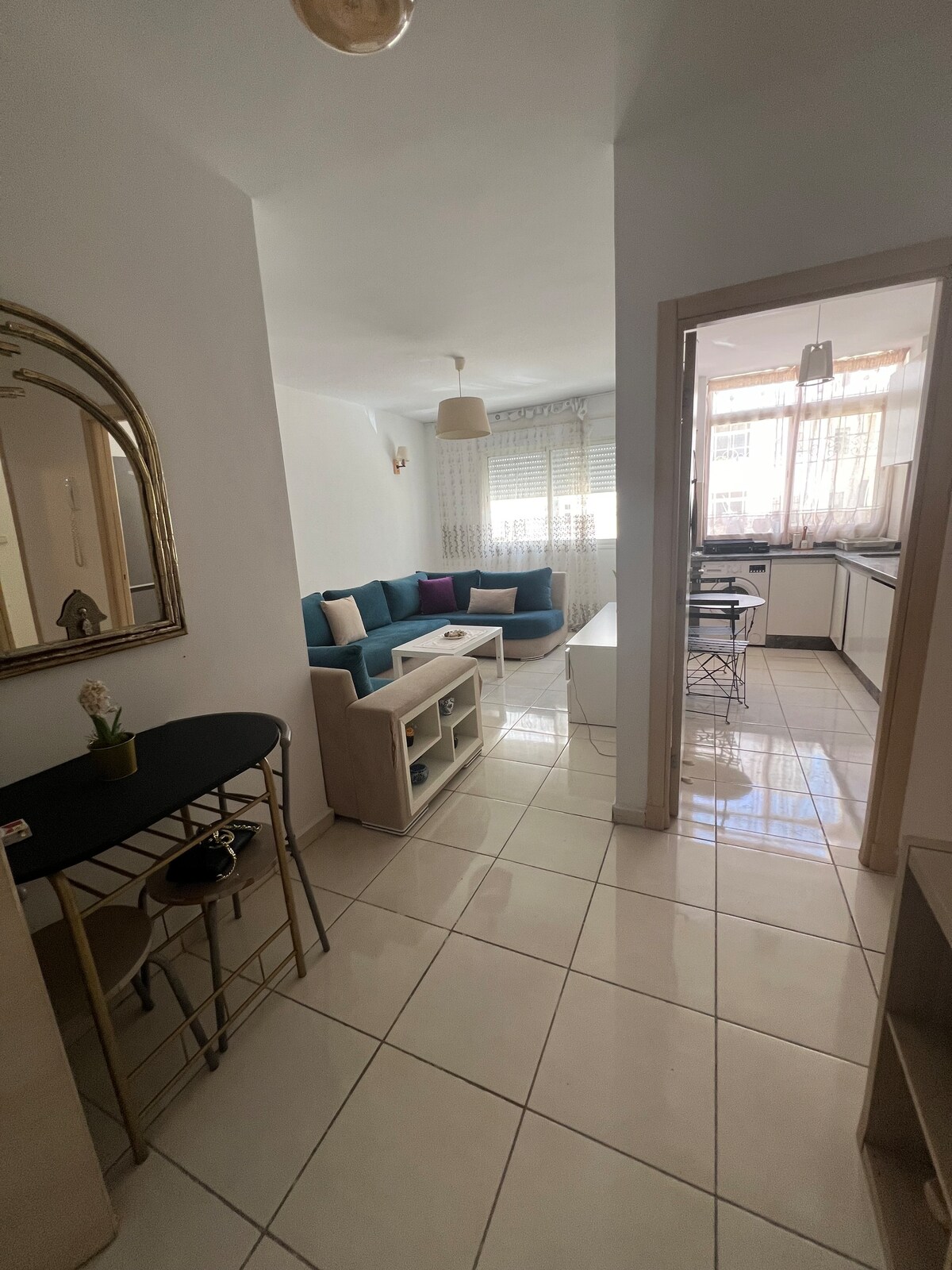 A bright and open living area features a blue sectional sofa with decorative pillows, alongside a white coffee table. Floor-to-ceiling windows allow natural light to fill the space. A dining table with chairs is visible, and a decorative mirror adds depth to the entryway.