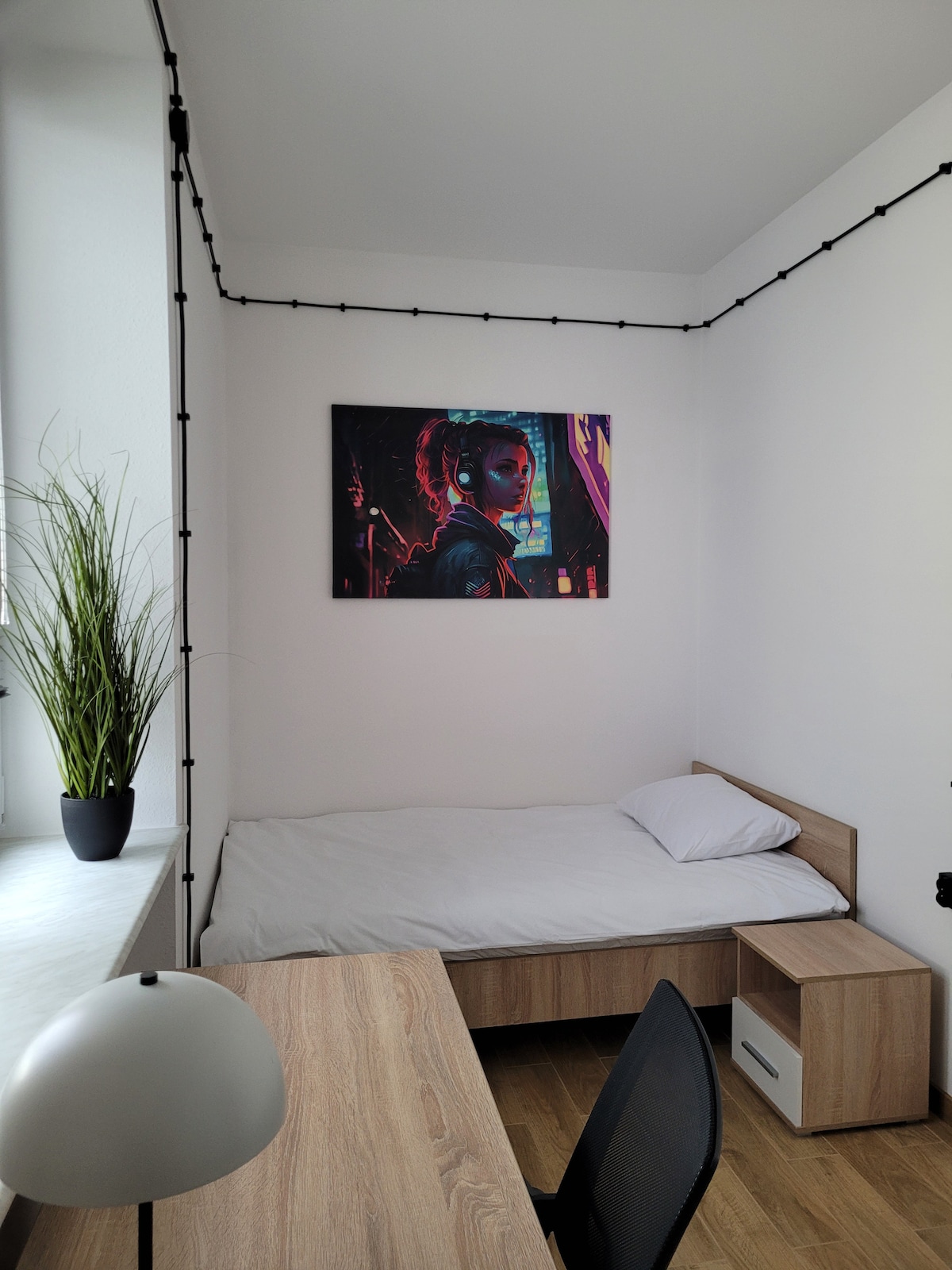 The room features a single bed with a simple white bedding set and a light-colored wooden frame. A small side table is positioned next to the bed, while a desk with a black chair is present. A potted plant adds greenery, and a vibrant art piece is displayed on the wall.